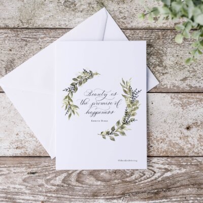 Beauty is the promise of happiness card staged "Beauty is the promise of happiness" note card with watercolor wreath. Staged on distressed wood background with eucalyptus leaves.
