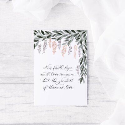 Now Faith Hope and Love Card staged "Now Faith Hope and Love" note card staged on distressed white wood background with eucalyptus leaves and white chiffon at the corner. Artwork features a watercolor wisteria with "now faith hope and love remain, but the greatest of these is love" in calligraphy