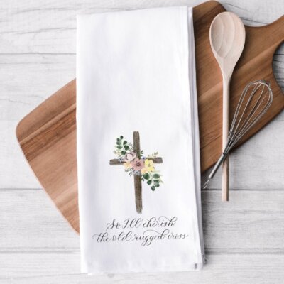 Old Rugged Cross kitchen towel staged on white tile background with wood cutting board, wire wisk, wooden spoon. Towel features a watercolor cross with flowers and "So i'll cherish the old rugged cross" in calligraphy beneath it.