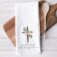 Old Rugged Cross kitchen towel staged on white tile background with wood cutting board, wire wisk, wooden spoon. Towel features a watercolor cross with flowers and "So i'll cherish the old rugged cross" in calligraphy beneath it.