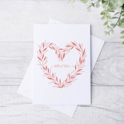 Pink "Mom" Mother's day card staged on a white wood background with eucalyptus. Artwork features pink wreath with "mom" in the center in calligraphy